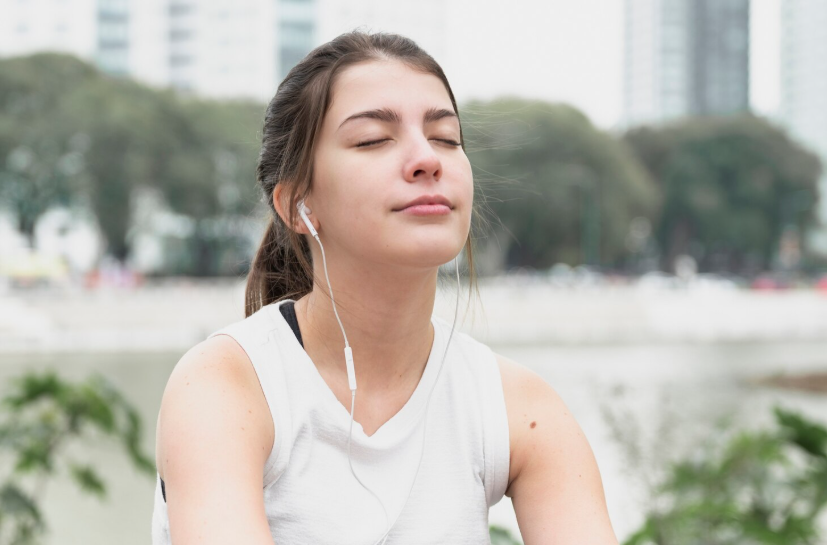 Woman with earbuds closed eyes and slow breath during a mindful breathing break in the park.