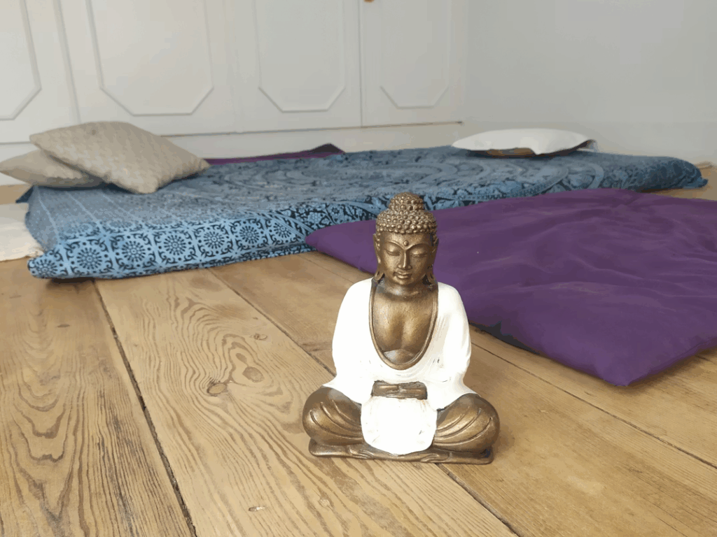 Small Buddha statue on wooden floor with cushions and mats arranged in a simple mindfulness corner at home.
