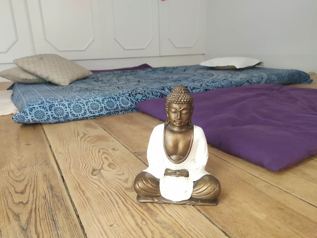 Small Buddha statue on wooden floor with cushions and mats arranged in a simple mindfulness corner at home.