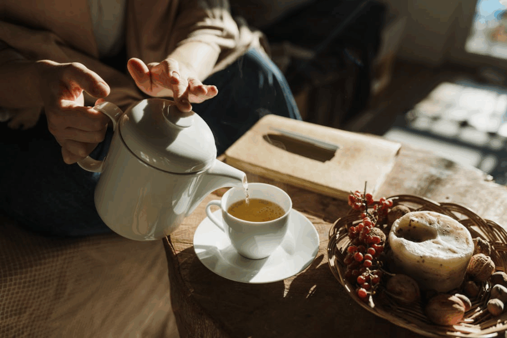 Hands pouring hot tea from a white teapot into a cup with candle and berries during a mindful tea ritual.