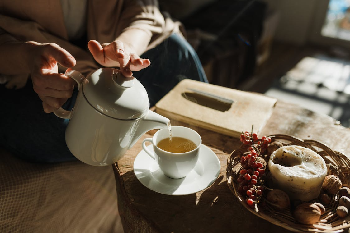 Hands pouring hot tea from a white teapot into a cup with candle and berries during a mindful tea ritual.