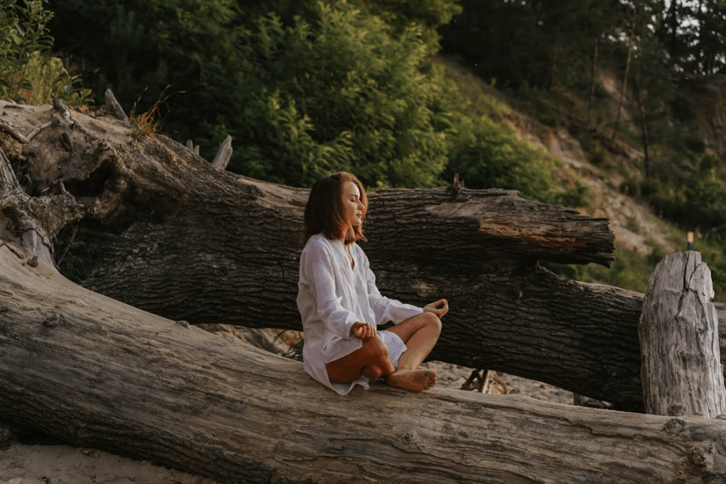 Woman sitting barefoot on a fallen tree, meditating in nature as she practices somatic grounding techniques.