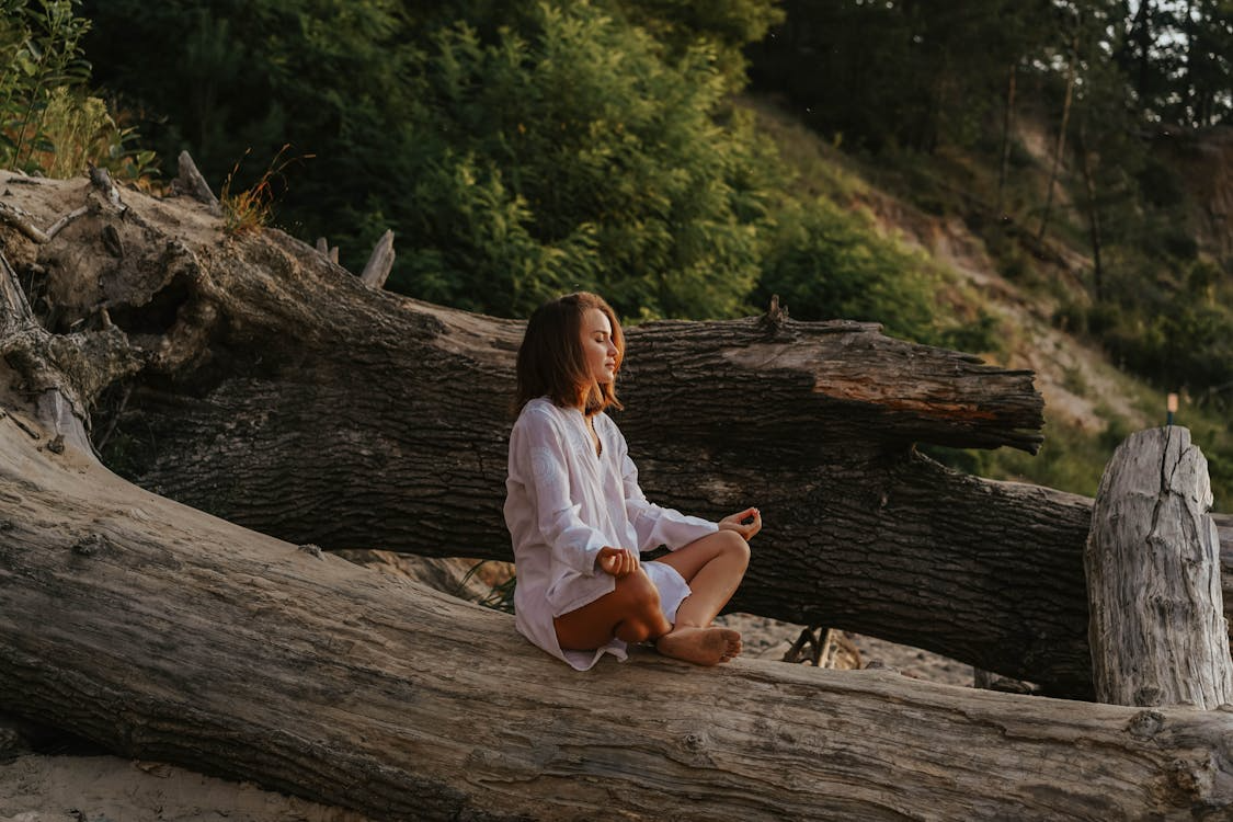 Woman sitting barefoot on a fallen tree, meditating in nature as she practices somatic grounding techniques.